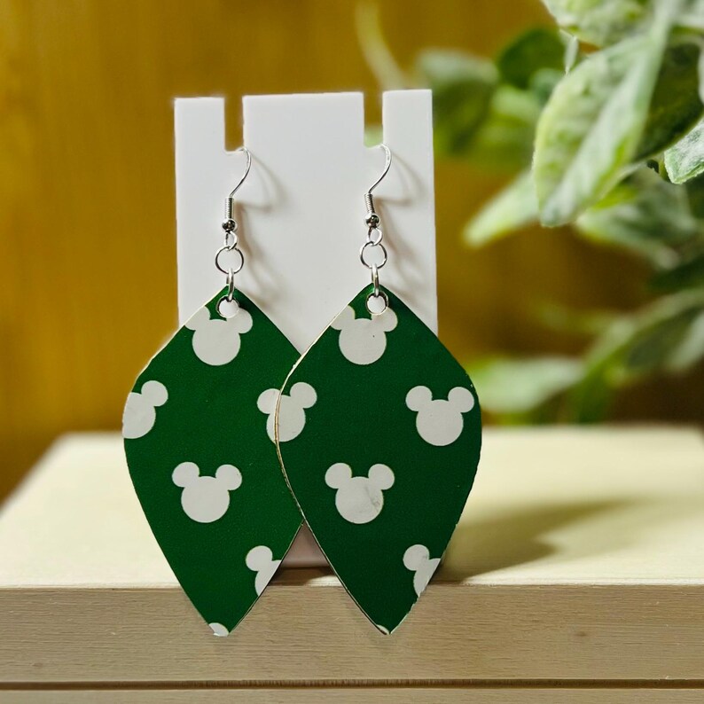 Puede incluir: Pendientes verdes en forma de l&aacute;grima con un patr&oacute;n de Mickey Mouse blanco. Los pendientes tienen ganchos plateados y se exhiben en un soporte blanco. El fondo incluye vegetaci&oacute;n y una superficie clara.