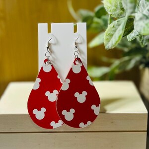 Puede incluir: Pendientes rojos en forma de l&aacute;grima con un patr&oacute;n de cabeza de Mickey Mouse blanca. Los pendientes cuelgan de ganchos plateados. Los pendientes se exhiben en un soporte blanco sobre un fondo borroso.