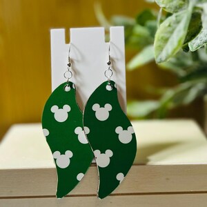 Puede incluir: Pendientes verdes en forma de hoja con un patr&oacute;n de Mickey Mouse blanco. Los pendientes cuelgan de ganchos plateados. Los pendientes se exhiben en un soporte blanco.