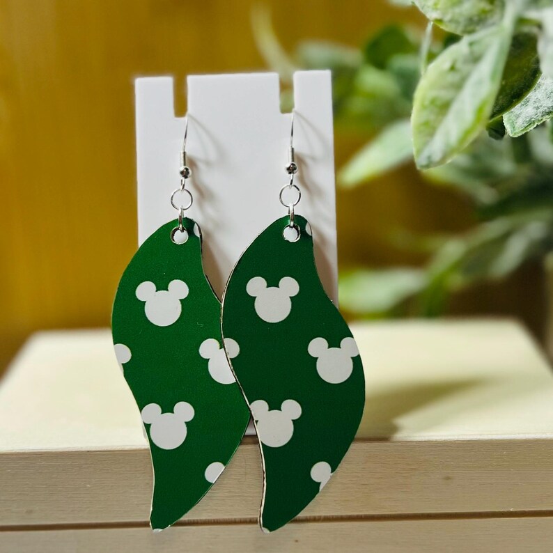 Puede incluir: Pendientes verdes en forma de hoja con un patr&oacute;n de Mickey Mouse blanco. Los pendientes cuelgan de ganchos plateados. Los pendientes se exhiben en un soporte blanco.