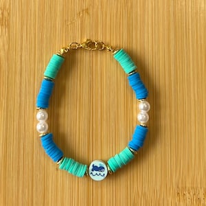 May include: A beaded bracelet featuring turquoise, blue, and gold accents. The bracelet includes a charm with a whale design and white pearl beads. The bracelet is on a wooden surface.