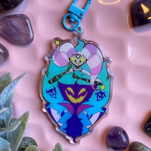 Puede incluir: Un personaje de dibujos animados azul y morado con un sombrero de bufón rosa y blanco. El personaje sostiene un corazón en su mano. El personaje está en un llavero de acrílico transparente con un cierre de metal azul.