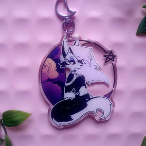 May include: A black and white anime-style fox character with pink accents sitting on a crescent moon with a star and floral design. The character has a white tail and a black body. The keychain is made of clear acrylic.
