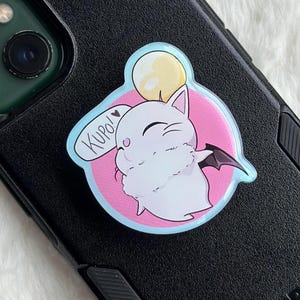 May include: A white cartoon character with a bat wing and a yellow orb, set against a pink circle, is affixed to a black phone case. The character has a speech bubble that says "Kupo!" in a playful font. The design is outlined in blue.