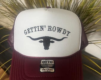May include: A burgundy and white trucker hat with the text "GETTIN' ROWDY" above a black longhorn silhouette. The hat has a mesh back and a foam front. The brand "OTTO" is visible on a tag.