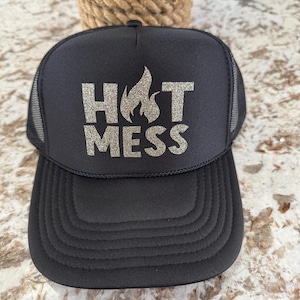 Puede incluir: Una gorra de camionero negra con un diseño de purpurina plateada que dice "HOT MESS".