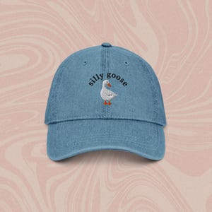 May include: A blue denim baseball cap with the text "silly goose" embroidered in black on the front. A white goose with orange feet is embroidered below the text.