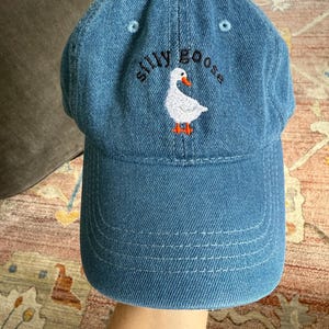 May include: A blue denim baseball cap with white stitching and an embroidered white goose with orange feet. The text "silly goose" is embroidered in black above the goose.