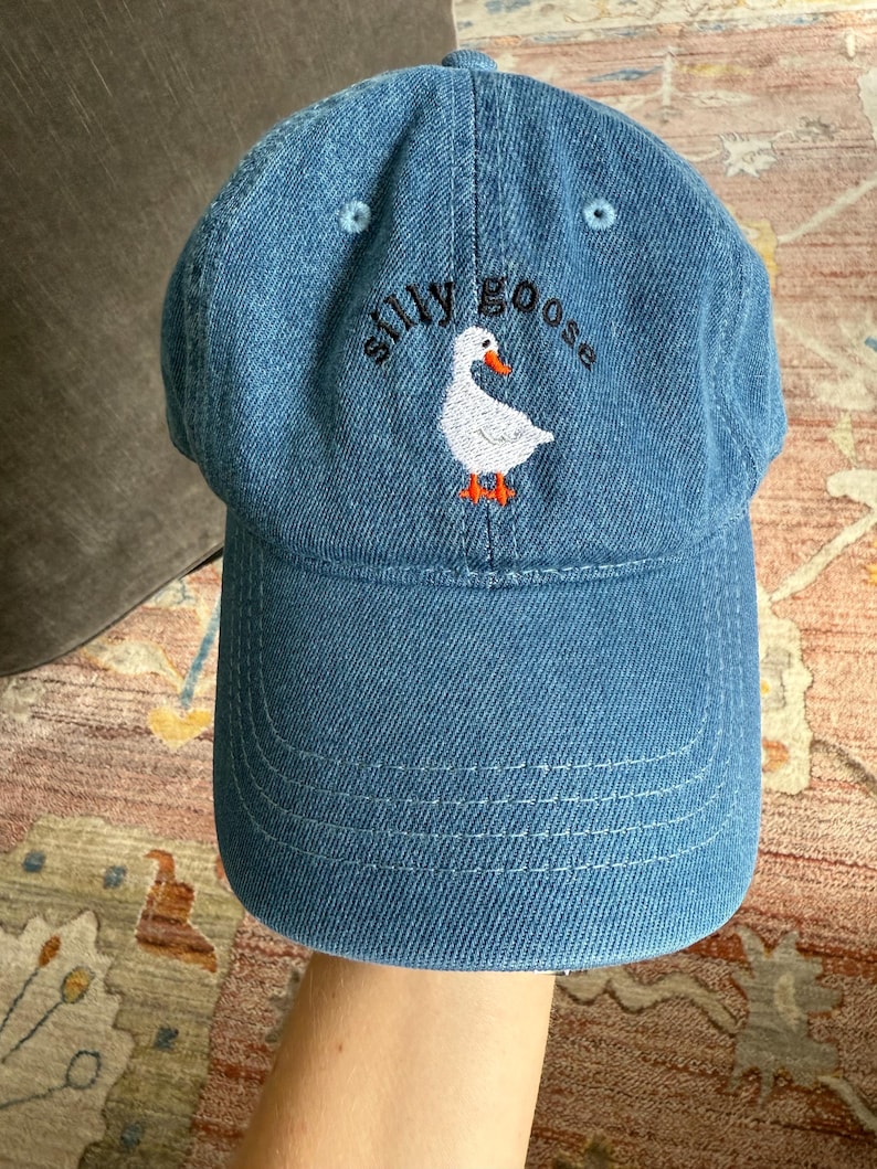 May include: A blue denim baseball cap with white stitching and an embroidered white goose with orange feet. The text "silly goose" is embroidered in black above the goose.