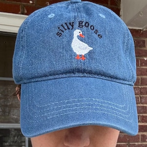 May include: A blue denim baseball cap with an embroidered white goose and the text "silly goose" in black.