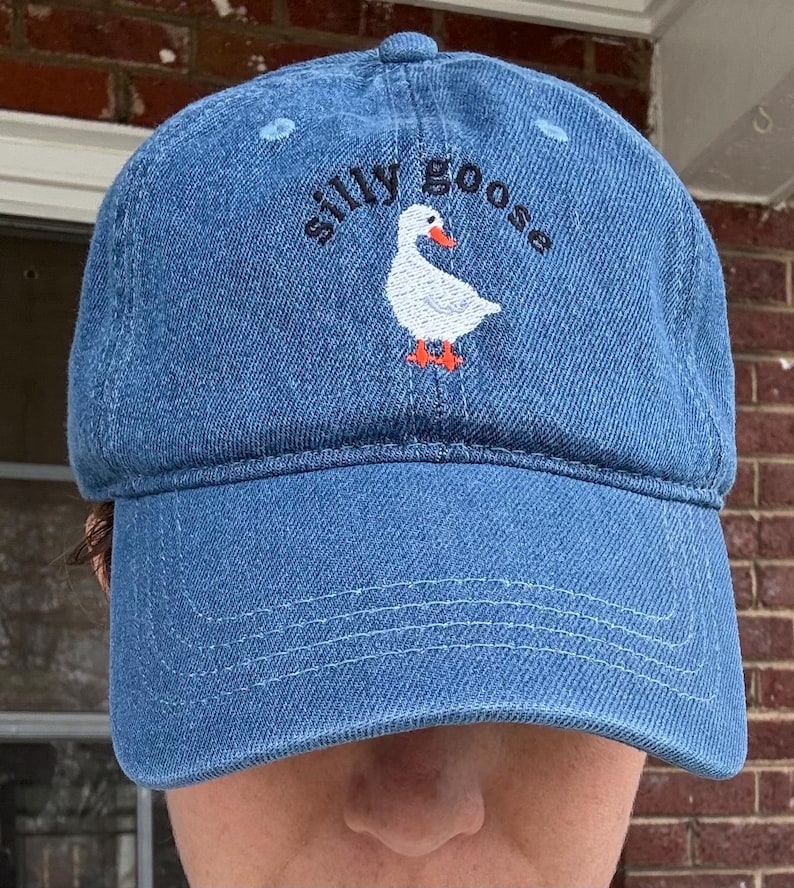 May include: A blue denim baseball cap with an embroidered white goose and the text "silly goose" in black.
