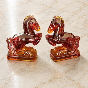 May include: A pair of amber-colored glass horse figurines, each depicted in a rearing pose on a rectangular base. The translucent glass features a deep reddish-brown hue with lighter golden tones, showcasing detailed manes and tails.