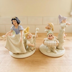 May include: Two porcelain figurines featuring Snow White in a blue and yellow dress with a dwarf playing a stringed instrument, and a second figurine with two dwarfs, one playing an accordion, all set on circular bases with gold trim accents.