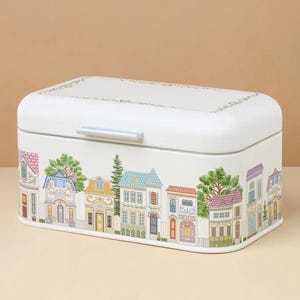 May include: White rectangular bread box with a lid and a silver handle. The box is decorated with a colorful illustration of a row of houses with trees and flowers. The houses have different architectural styles and colors.