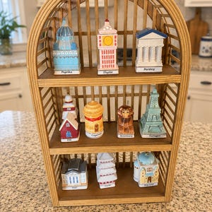 May include: A decorative bamboo shelf displays a collection of miniature spice jars shaped like landmarks. The jars are painted in various colors and feature labels such as "Paprika," "Thyme," and "Parsley." The shelf has a curved top and multiple wooden shelves.