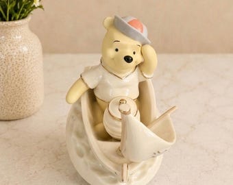 May include: A ceramic figurine of a yellow bear wearing a white shirt and a grey and pink hat, sitting inside a small cream-colored boat. The bear holds a small honey pot, and the boat features a white sail and a textured, dimpled exterior surface.