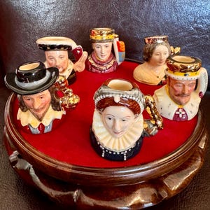 Puede incluir: Conjunto de siete tazas de cerámica en miniatura con retratos de figuras históricas, incluyendo un rey, una reina y otros miembros de la realeza. Las tazas se muestran en una base de madera con un forro de terciopelo rojo.