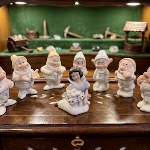 May include: A collection of eight porcelain figurines featuring a central female character with black hair and a blue bodice, surrounded by seven small male figures wearing hats and white clothing, all arranged on a polished wooden surface.