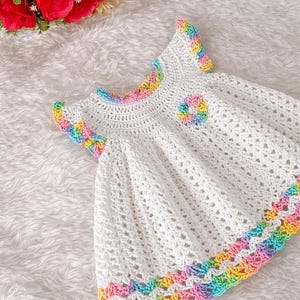 Close-up of a white crocheted baby dress featuring rainbow scalloped trim on sleeves, collar, and hem.