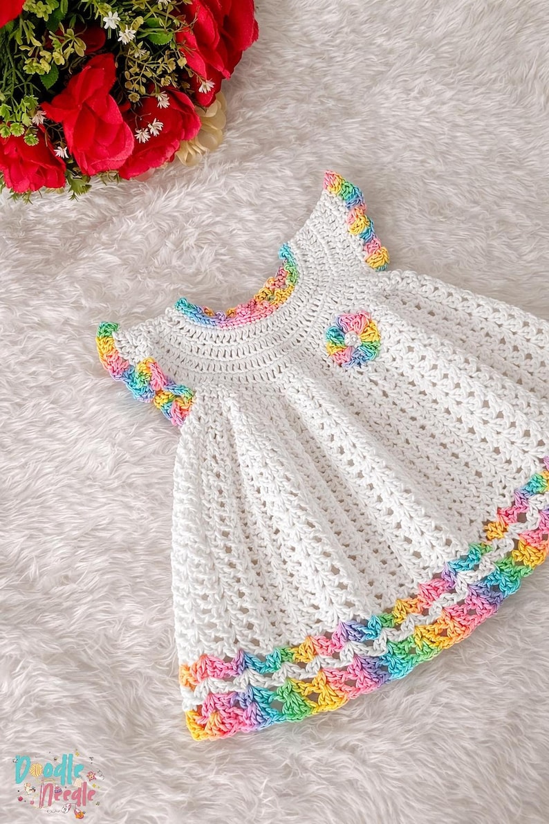 Close-up of a white crocheted baby dress featuring rainbow scalloped trim on sleeves, collar, and hem.
