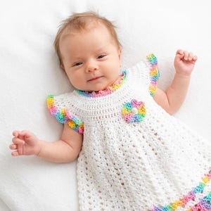A baby lying on a white surface wearing a handmade white dress with colorful crochet accents.
