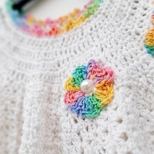 Detailed close-up shows the texture of the white crocheted bodice and a small rainbow flower applique with a pearl center.