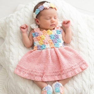 Puede incluir: Un vestido de bebé tejido a ganchillo con un corpiño de motivos florales en tonos pastel multicolor con centros blancos, una falda rosa claro, una diadema floral a juego y un par de patucos blancos tejidos con detalles florales pastel.