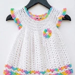 A white crocheted baby dress with rainbow edges hangs from a black hanger against a plain white wall.