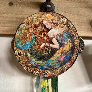 Peut inclure: Une décoration ronde en bois avec une illustration colorée de sirène. La sirène a de longs cheveux blonds flottants et une queue bleue et verte. La décoration est décorée de volutes dorées et est suspendue à un crochet.