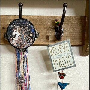 Puede incluir: Una estantería de madera con dos ganchos. En el gancho izquierdo cuelga un pandero redondo blanco y negro con un diseño de unicornio. En el gancho derecho cuelga un cartel que dice "Cree en tu propia magia" con una cuerda de pájaros coloridos colgando debajo.