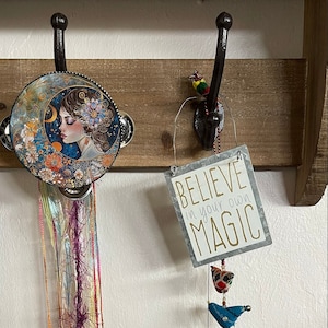 May include: A wooden shelf with two hooks. On the left hook hangs a tambourine with a colorful illustration of a woman's face. On the right hook hangs a sign that reads "Believe in your own magic" with a colorful bird mobile hanging below.