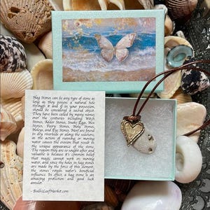 May include: A natural hag stone pendant with a gold-colored heart charm, displayed in a small gift box with a beach scene. The pendant hangs on a brown cord, surrounded by seashells. Text on the box describes the stone's properties.