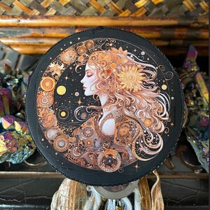 Puede incluir: Una caja redonda decorativa negra con una imagen dorada y blanca de una mujer con el pelo largo y una corona de girasol. La imagen está rodeada de intrincados detalles de engranajes y cuerpos celestes.