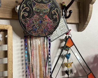May include: A black tambourine with a colorful image of a cat with intricate details. The tambourine is hanging from a hook with colorful yarn streamers.