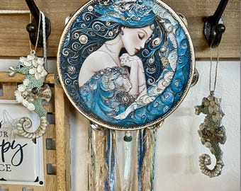 May include: A round wall hanging featuring a serene woman with long blue hair, adorned with pearls and a flowing blue gown. Two seahorse ornaments hang on either side. A wind chime with blue and beige strands hangs below the wall hanging.