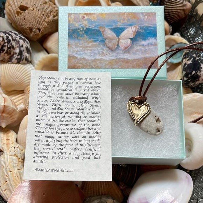 May include: A natural stone pendant with a gold-colored heart charm, displayed in a small box with a butterfly image. The pendant hangs on a brown cord, surrounded by seashells. Text on a card describes hag stones.