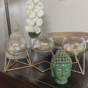 May include: Three clear glass bowls with iridescent finishes, each resting on a gold-toned geometric metal stand. The bowls are filled with various small stones and shells. A green ceramic Buddha head sculpture sits in front of the stands.