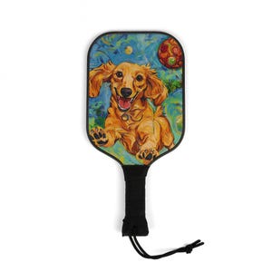 May include: Pickleball paddle featuring a happy dachshund dog illustration. The paddle has a black handle with a wrist strap. The background is a blue and green abstract design with a red and orange ball.
