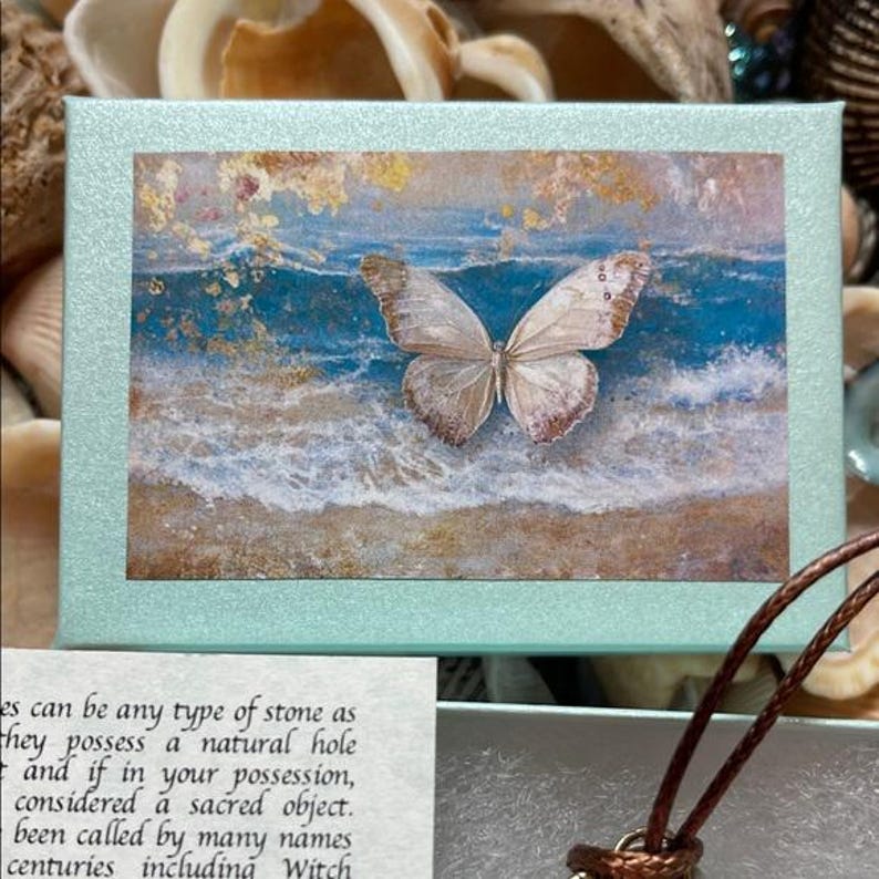 May include: A light blue box with a watercolor painting of a butterfly over a beach scene. The butterfly is light brown with darker brown markings. The box is surrounded by seashells and a brown leather cord.