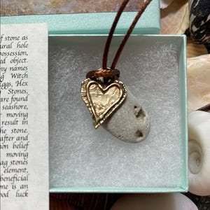 May include: A heart-shaped pendant with a textured gold finish is attached to a gray stone. The pendant hangs from a brown cord, presented in a light blue box. The stone has natural holes and is a unique piece of jewelry.