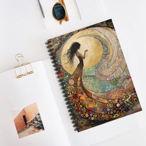 Op de afbeelding: Spiraalgebonden notitieboek met een kleurrijke illustratie van een vrouw met lang, golvend haar en een patroonjurk. Het kunstwerk bevat een grote gele maan en wervelende, abstracte ontwerpen in verschillende kleuren. Het notitieboek ligt open op een witte ondergrond.