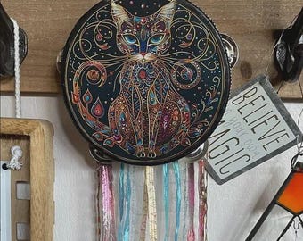 May include: A round, black tambourine with a detailed, colorful cat design. The cat is depicted with intricate patterns and jewel-toned accents. Below the tambourine are colorful ribbons. A sign that says "BELIEVE YOUR OWN MAGIC" is visible.