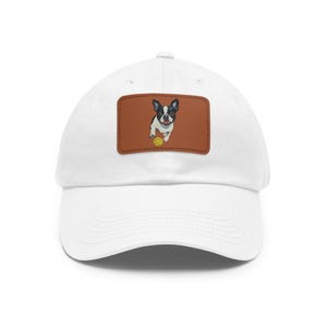May include: White baseball cap with a brown leather patch showing a Boston Terrier dog playing with a yellow ball. The dog is black and white. The cap has a curved peak and a classic, casual style.