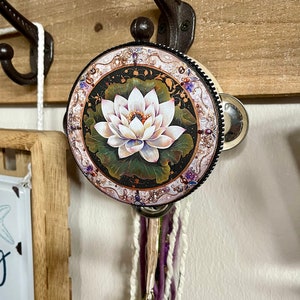 May include: A white and purple tambourine with a floral design featuring a white lotus flower on a black background. The tambourine is hanging from a hook on a wooden wall.