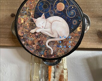 May include: A white cat sleeps on a crescent moon against a dark blue background with gold swirls. The moon is part of a round, black tambourine with a silver jingle.