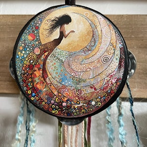 May include: A round, colourful tambourine with a black handle and a painted image of a woman with flowing hair and a long, flowing dress. The tambourine is decorated with a variety of colourful dots, swirls, and patterns.
