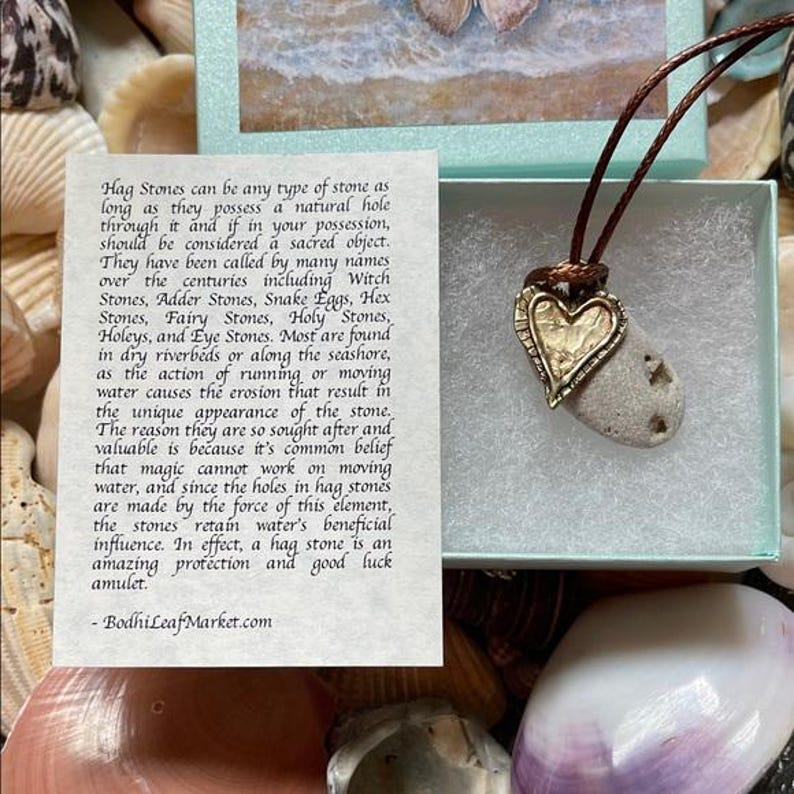 May include: A natural hag stone pendant with a heart-shaped gold-colored metal accent. The stone is attached to a brown cord necklace. The pendant is displayed in a gift box with a seashell background. Text on a card describes hag stones.