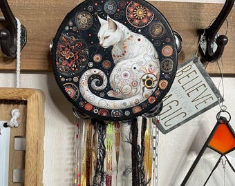 May include: A round, black wind chime featuring a white cat with intricate, colorful patterns. The cat is set against a backdrop of celestial designs. The chime hangs from a wooden surface with decorative hooks and a metal sign that reads "BELIEVE YOUR OWN MAGIC."