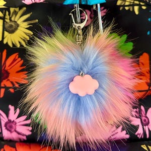 May include: A fluffy, multi-colored faux fur pom-pom keychain featuring a soft pastel pink cloud charm attached to a silver-toned metal lobster clasp and ring. The pom-pom displays a blend of light blue, pink, and yellow hues throughout its fibers.