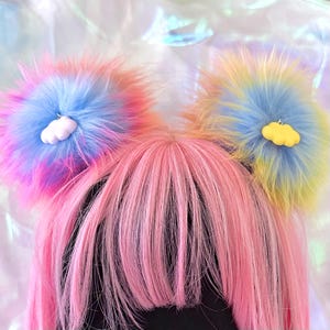 May include: A pair of fluffy, multi-colored faux fur pom-pom hair accessories featuring a tie-dye pattern of pink, blue, and yellow. Each pom-pom is accented with a small, solid-colored cloud charm, one in white and one in yellow, attached to the center.